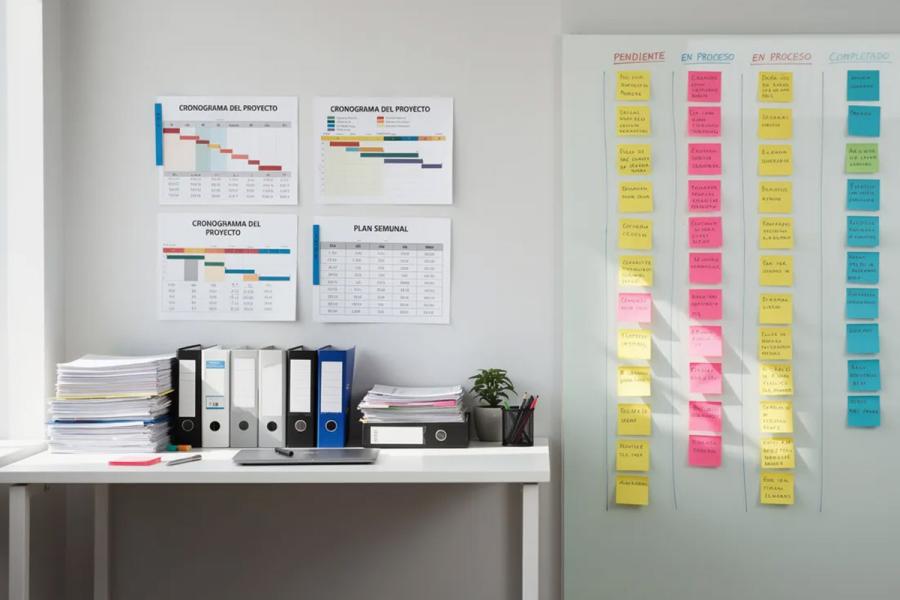 El espacio de trabajo está dividido en dos secciones: a la izquierda, hay documentos y cronogramas impresos relacionados con la gestión de proyectos, mientras que a la derecha se encuentran pizarras llenas de notas adhesivas de colores organizadas en columnas, representando diferentes tareas y fases del ciclo de vida del proyecto.